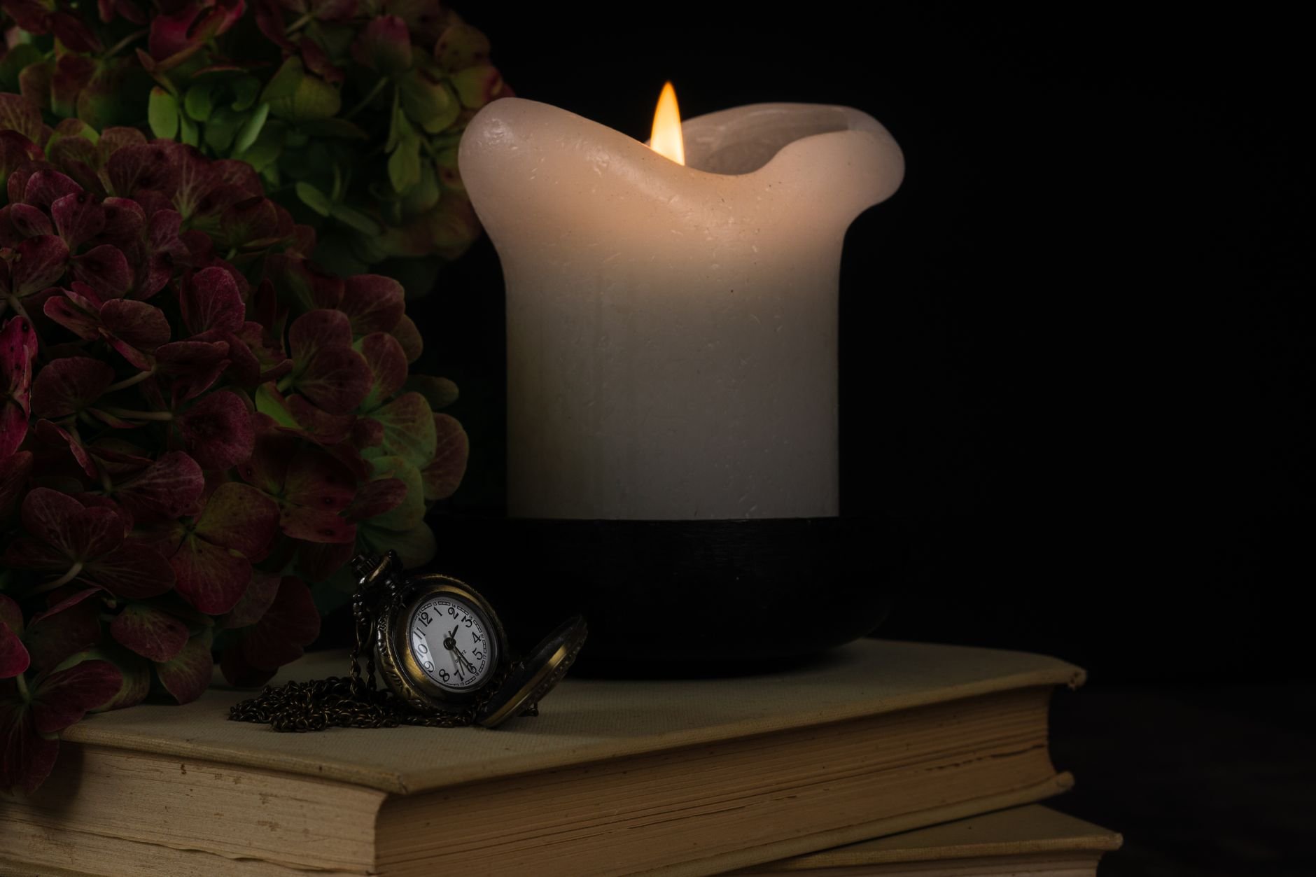 A serene still life featuring a lit candle, antique pocket watch, and books — warm candlelight on dark surface.